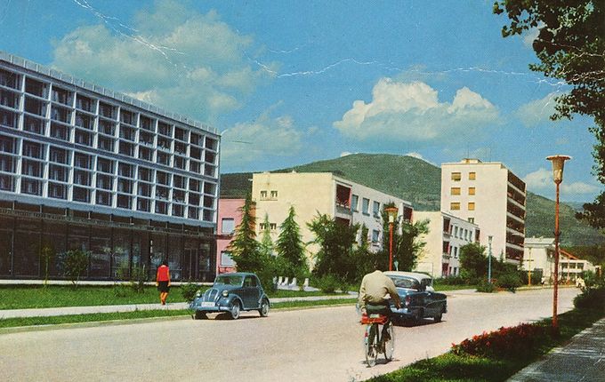 Erjavčeva ulica ob koncu 60. letih. Od leve proti desni: sedež Elektro Primorska, sedež Soških elektrarn, prvi »nebotičnik«, v katerem je bila prva (in še vedno delujoča) samopostrežna trgovina v mestu. V ozadju na sredini hrib Škabrijel. Vir razglednice: Pokrajinski arhiv Nova Gorica.  